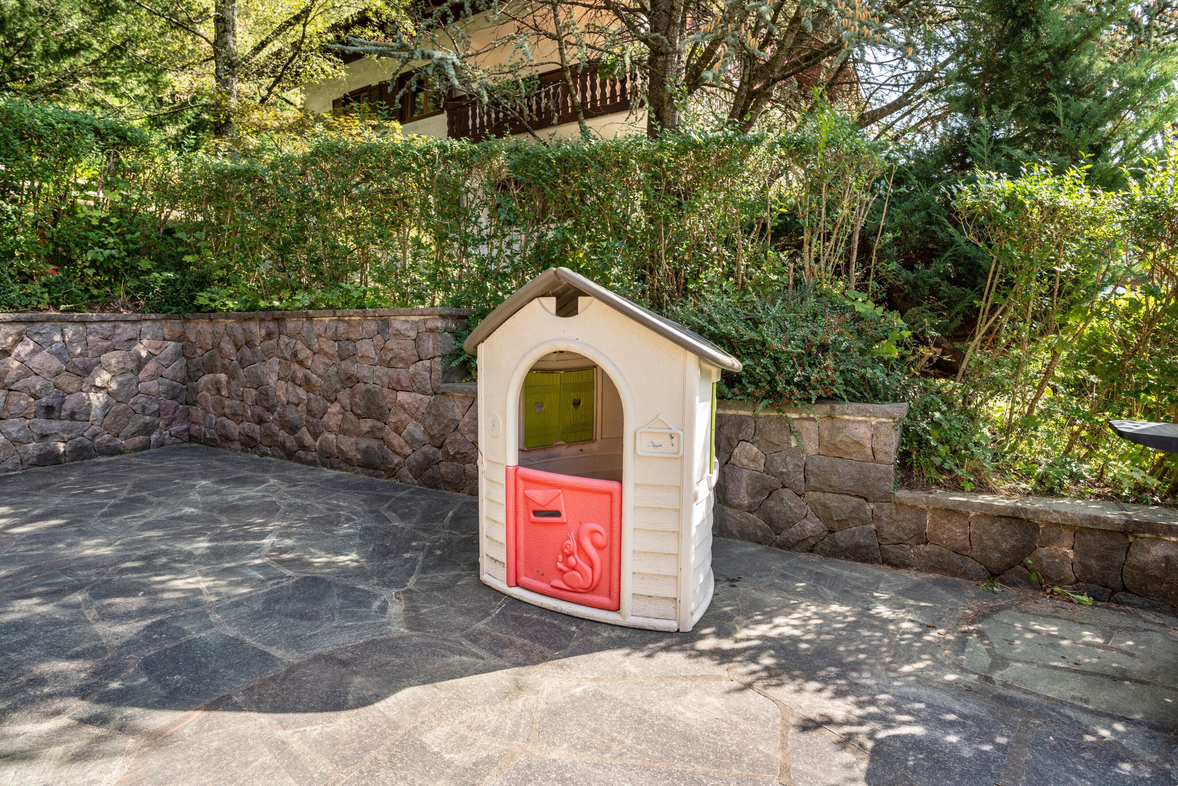 A small, stone-walled playhouse with a red mailbox, surrounded by greenery.