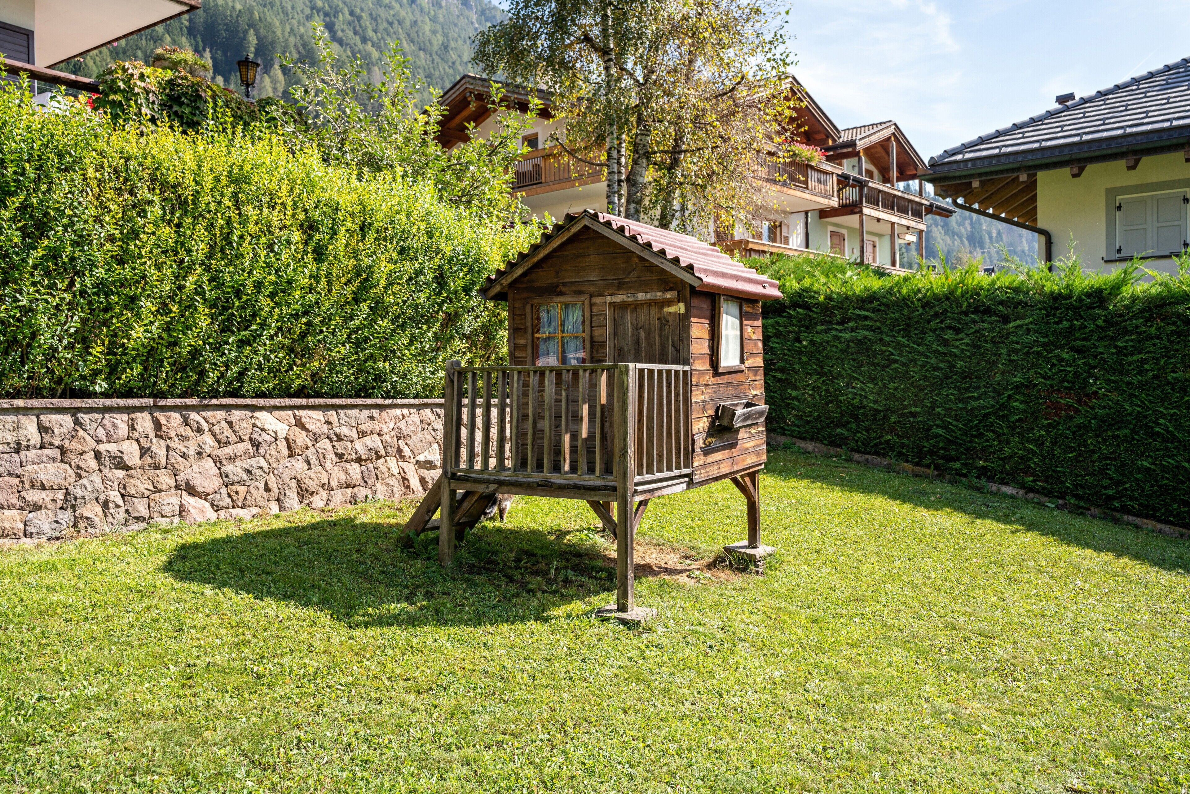 A wooden playhouse in a grassy area with a stone wall and a hedge in the background.