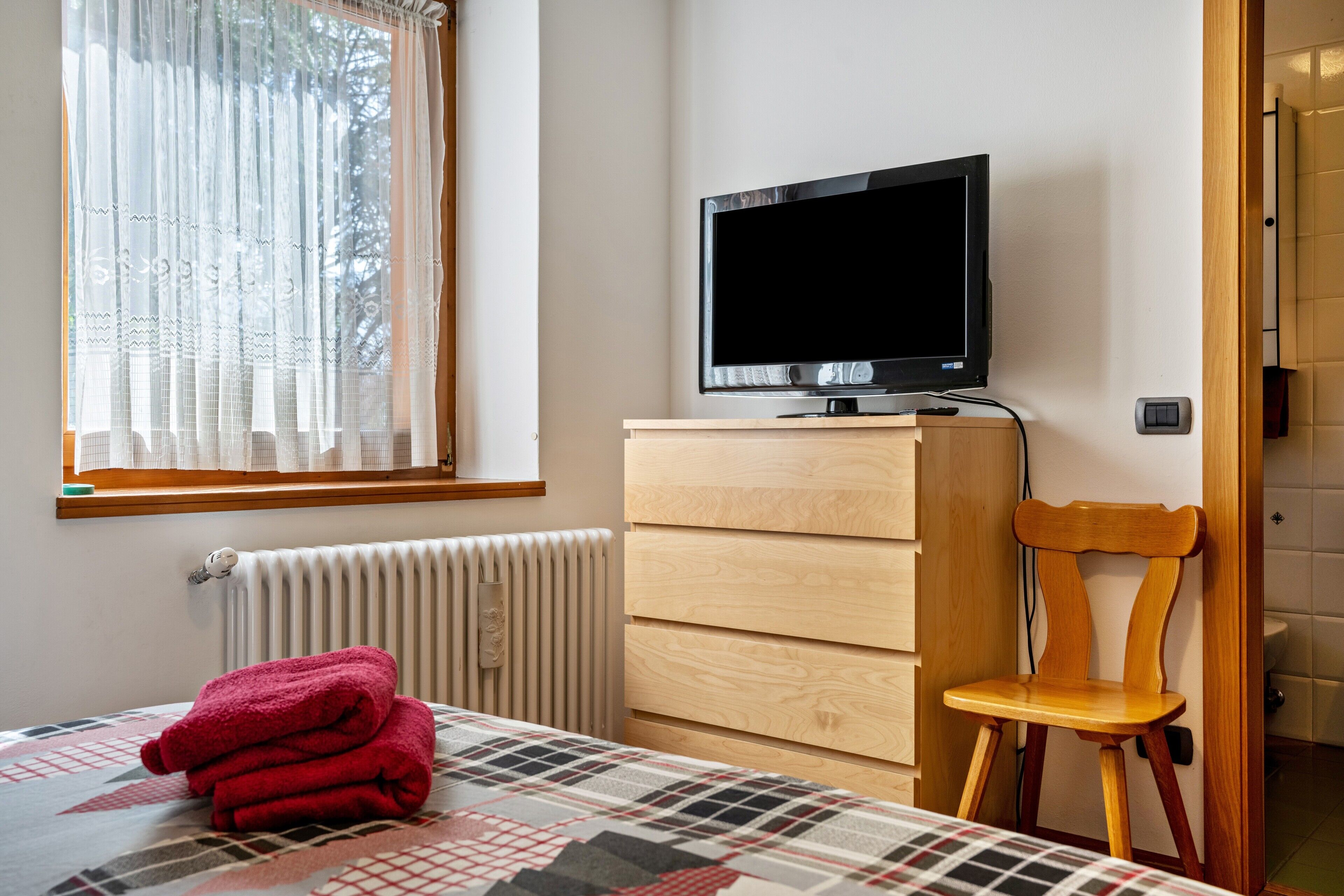 A bedroom with a bed, a TV on a wooden dresser, a chair, and a window with curtains.