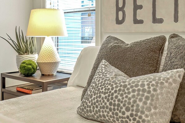 Un dormitorio ordenado con cama, almohadas, una lámpara, una planta en maceta y un libro en la mesita de noche.