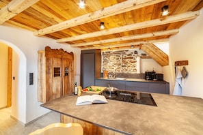 A kitchen with a stone wall backsplash, wooden ceiling beams, and a central island with a built-in stove.