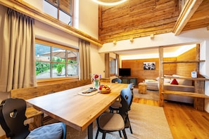 A wooden dining table with chairs, a television, and a sofa in a room with wooden beams and large windows.
