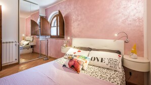 Ein Schlafzimmer mit einem Bett, einem Nachttisch, einem Spiegel und einem Fenster mit Holzrahmen.