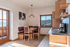 A modern kitchen with wooden cabinets, a dining area with a wooden table and chairs, a pendant light, and a view through a window.