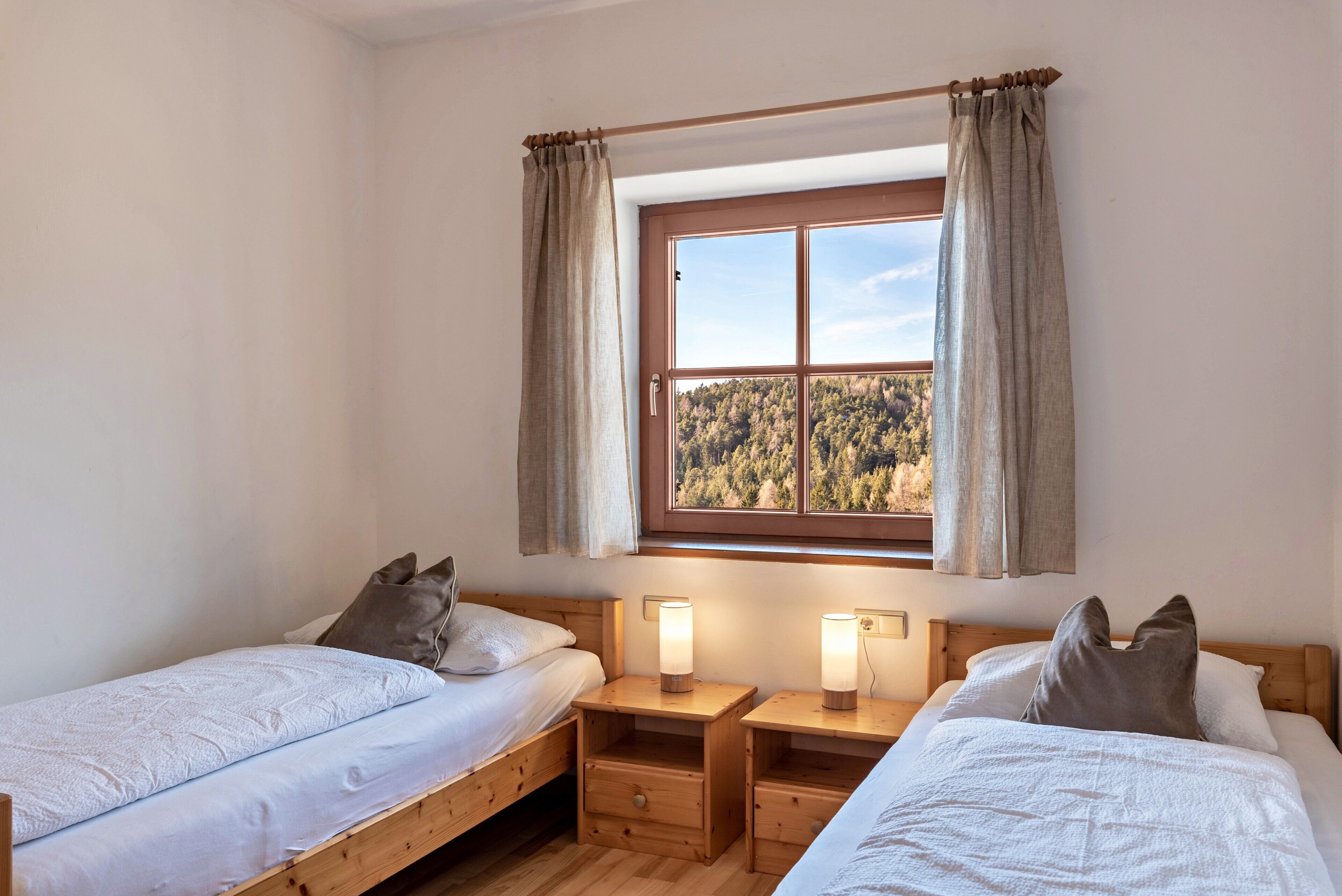 Ein Zimmer mit zwei hölzernen Betten, einem Fenster mit Vorhängen und Nachttischlampen.