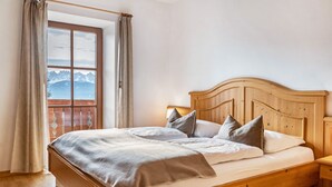Ein hölzernes Bett mit Kopfteil, ein Fenster mit Vorhängen und Blick auf Berge.