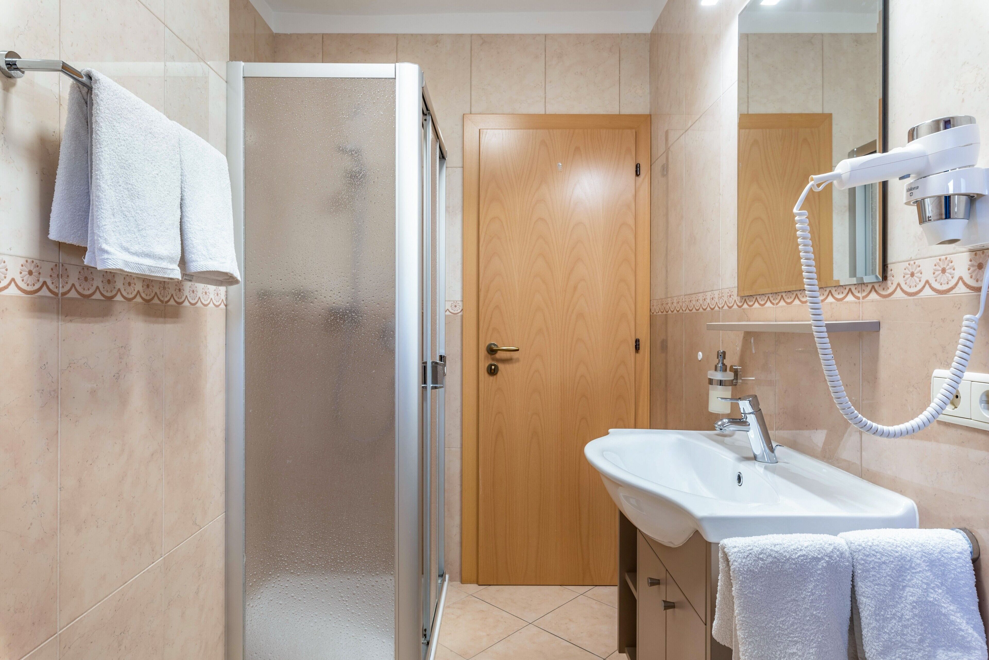 Un baño con mampara de vidrio, puerta de madera, lavabo blanco y toallero.