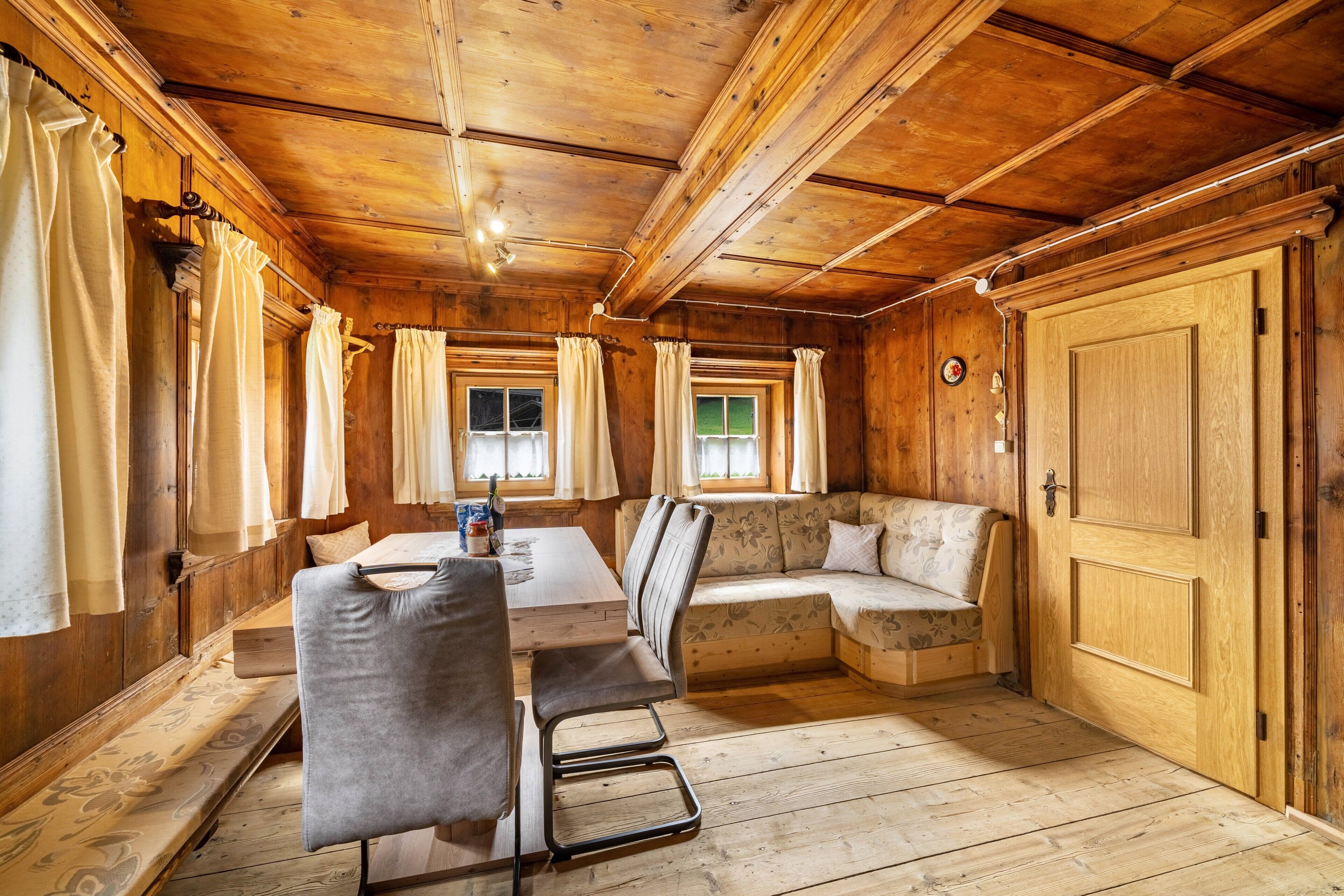 A wooden cabin interior with a dining table, chairs, a sofa, and a large wooden door.