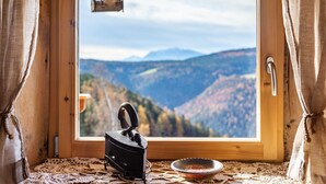 L’intérieur d’un chalet rustique, avec une fenêtre en bois, un poêle en fer noir et un chemin de table en tissu tressé posé sur le rebord de la fenêtre.