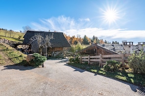 Un paysage rural avec un bâtiment à toit de chaume, une clôture en bois et un chemin de gravier menant à une maison dont le toit est en pente.