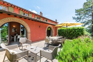 An outdoor patio with tables and chairs, a yellow umbrella, and a red building with arches. - Apartment In A Hotel "Villa Pesca" with Shared Pool, Private Balcony & Wi-Fi (Trequanda)