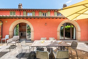 An outdoor patio with a table and chairs, a yellow umbrella, and a red building with arched doorways. - Apartment In A Hotel "Villa Pesca" with Shared Pool, Private Balcony & Wi-Fi (Trequanda)