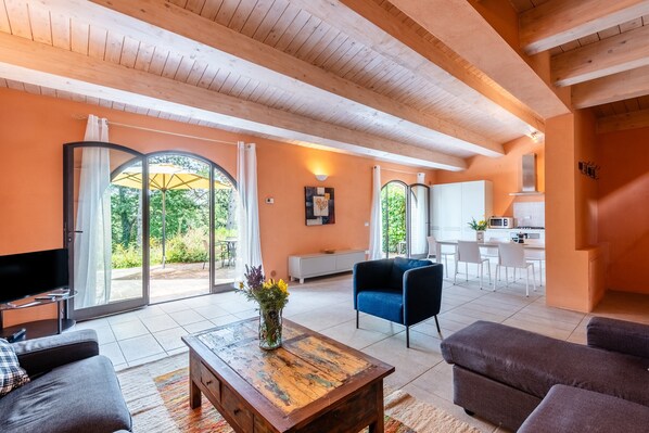 A modern living room with a television, sofa, coffee table, and a dining area with a kitchen in the background. - Apartment In A Hotel "Villa Pesca" with Shared Pool, Private Balcony & Wi-Fi (Trequanda)