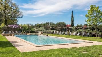 A rectangular pool with a stone border, surrounded by a grassy lawn and a pergola with chairs.