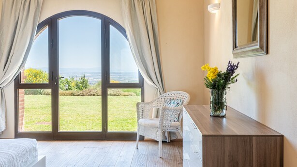 A room with a large arched window, a wicker chair, a wooden desk, and a vase with flowers.