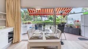 Un comedor con una mesa dispuesta para dos, una cocina con armarios blancos y vistas a un jardín por un cerramiento de vidrio.