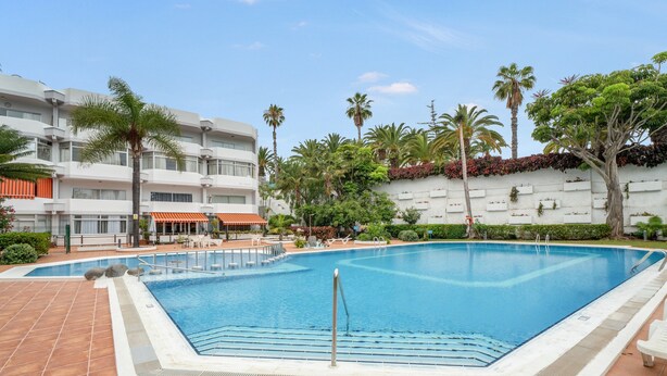 Una piscina con cubierta de baldosas, rodeada de palmeras y un edificio blanco con balcones.