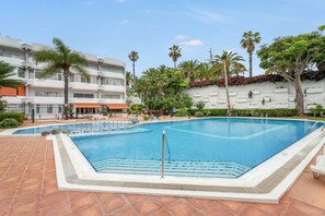 Una piscina con cubierta de baldosas, rodeada de palmeras y un edificio blanco con balcones.