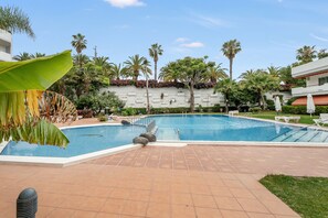 A swimming pool with a tiled deck, surrounded by palm trees and a white building with a red roof.