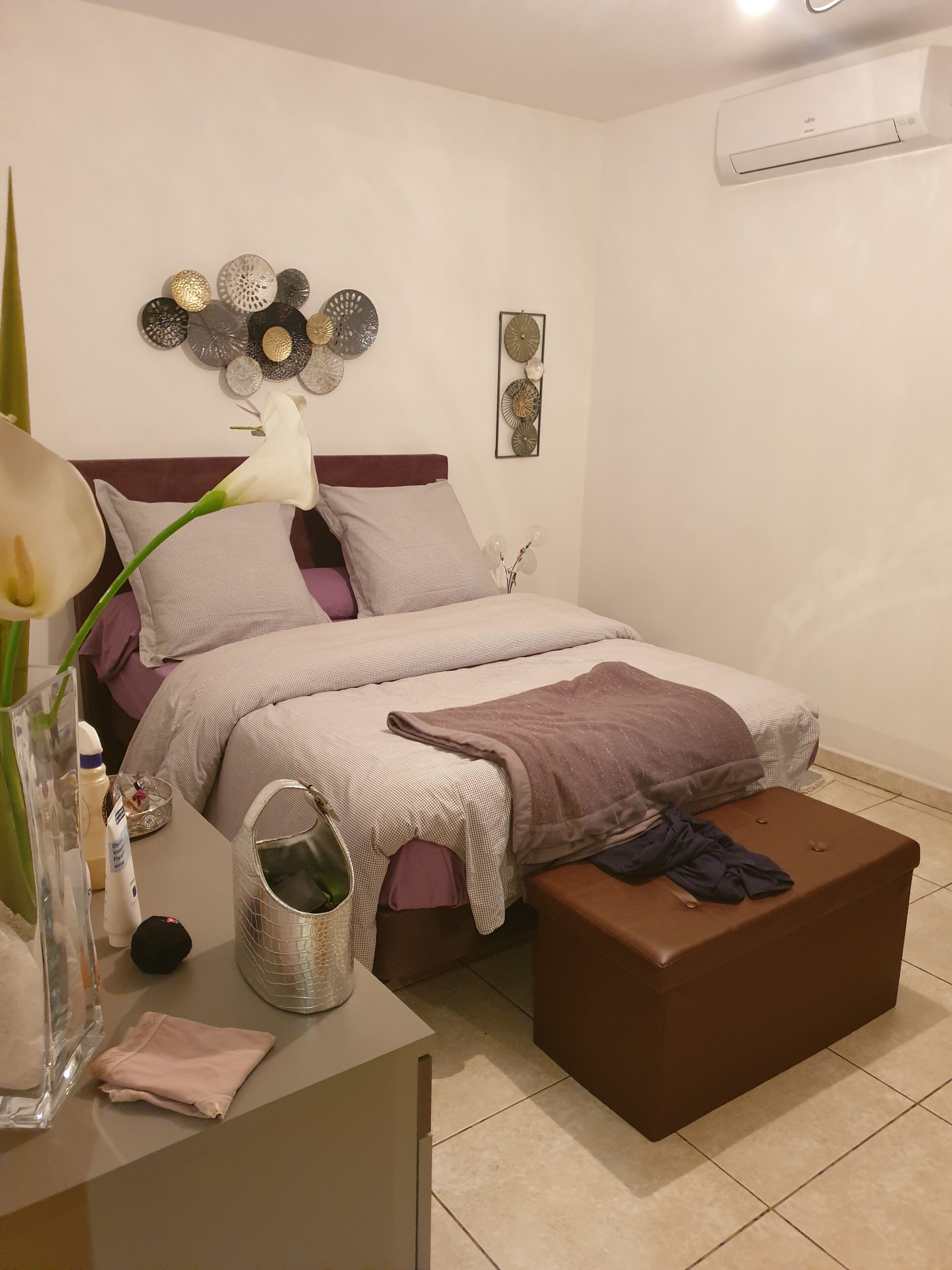 A bedroom with a bed, bedside table, and a brown ottoman.