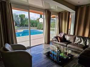 A living room with a glass coffee table, a leather sofa, and a view of a swimming pool through large sliding glass doors.