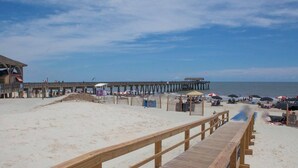 A beach with a wooden pier extending into the water, a sandy beach area, and beach umbrellas. - Pier Walk B ~ 2 Kings, 1 Queen, 2 Twins, 3 cars (Tybee Island)