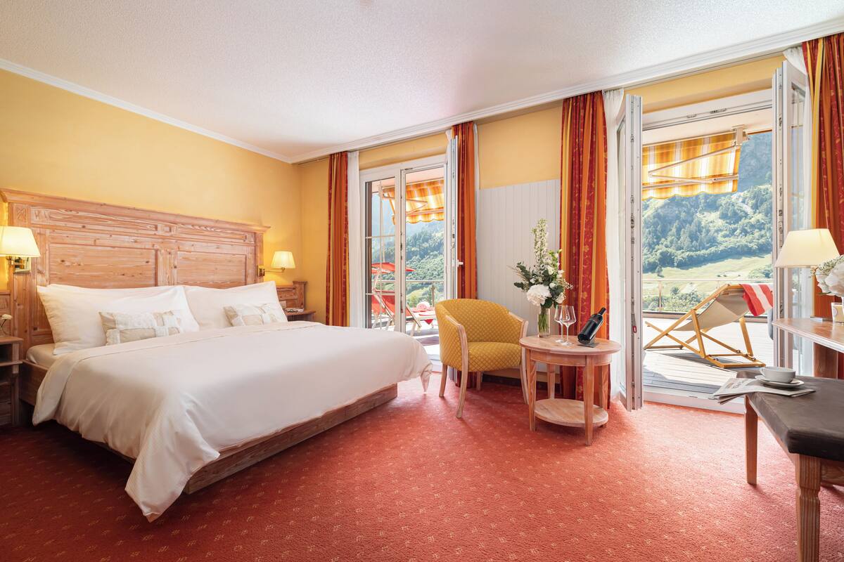 Habitación de hotel con una cama grande, cabecera de madera, un sillón amarillo, una mesita con un florero y vistas a un balcón con una silla.