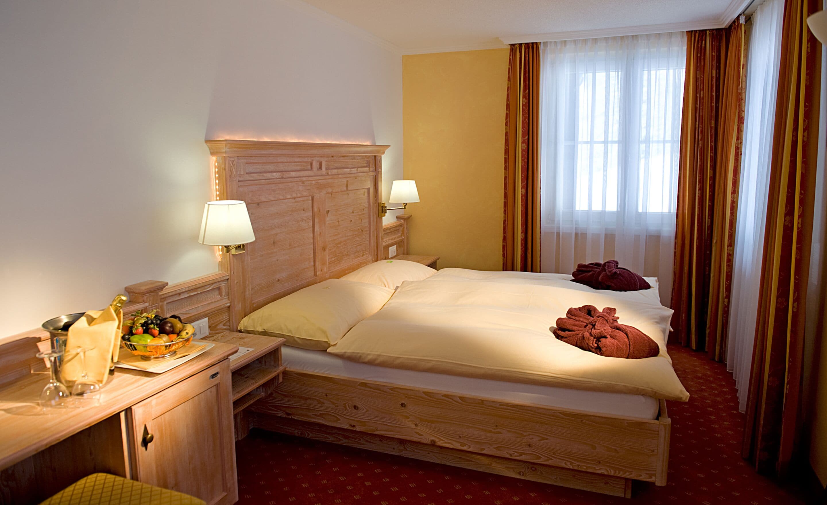 A hotel room with a wooden bed, two bedside lamps, a nightstand with a fruit bowl, and a red carpeted floor.