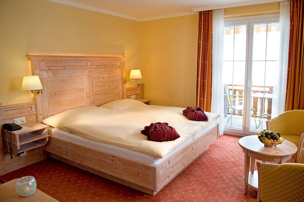 Habitación de hotel con cabecera de madera, dos lámparas de noche, cama con ropa blanca, una mesita redonda con fruta y un balcón con sillas.