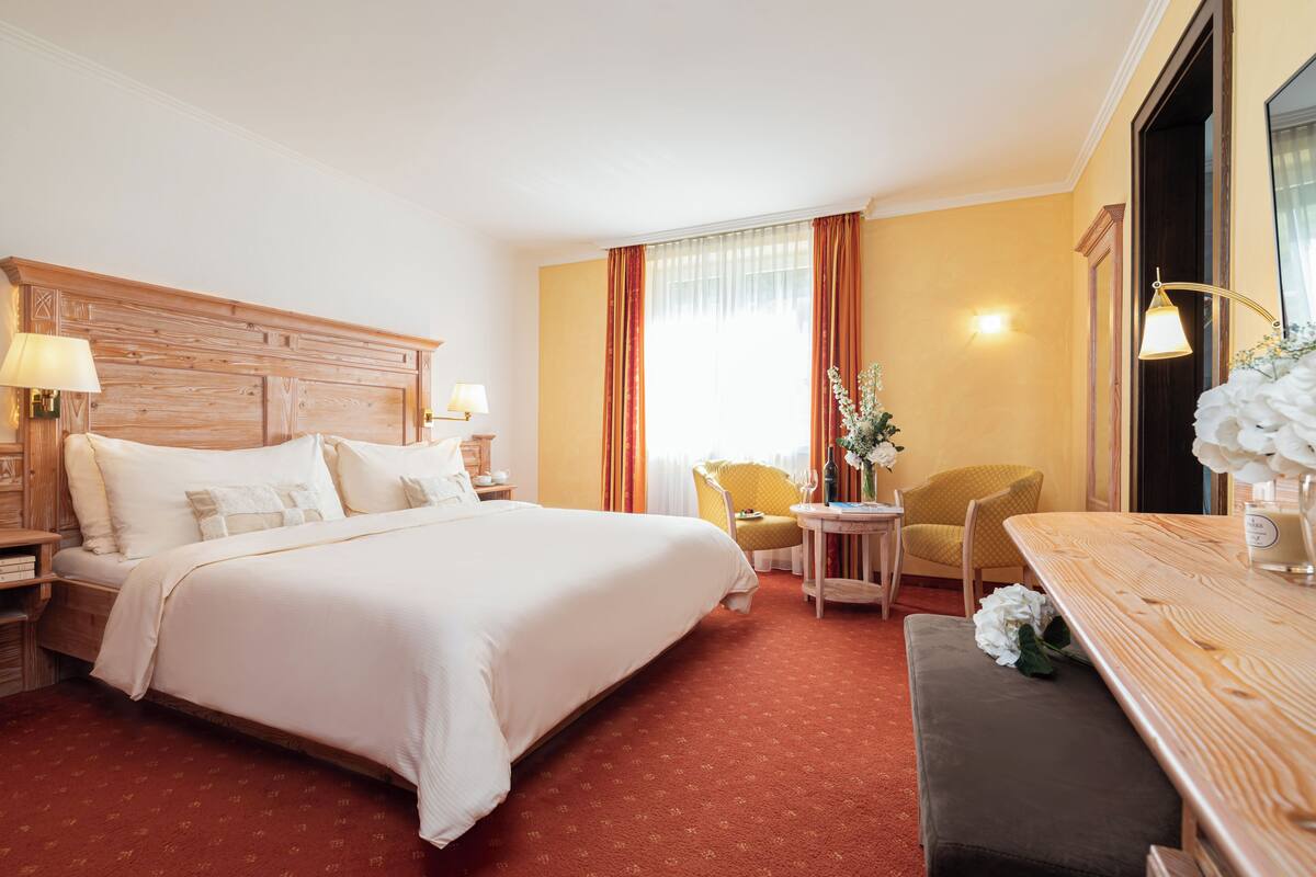 Habitación de hotel con una cama grande, cabecero de madera, una mesa redonda pequeña con una silla, un banco y una lámpara de pared.