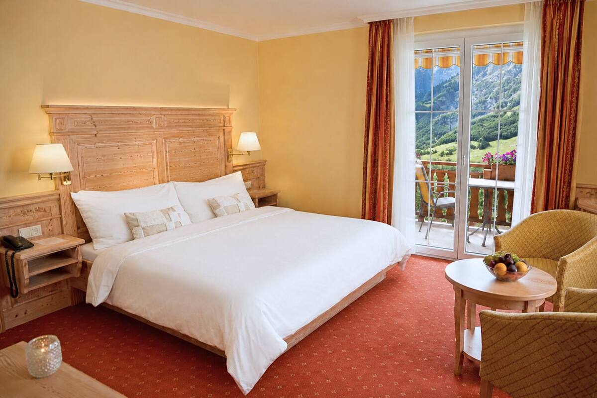 Habitación de hotel con una cama grande, dos lámparas de noche, una mesita con fruta, una silla y un balcón con vistas.