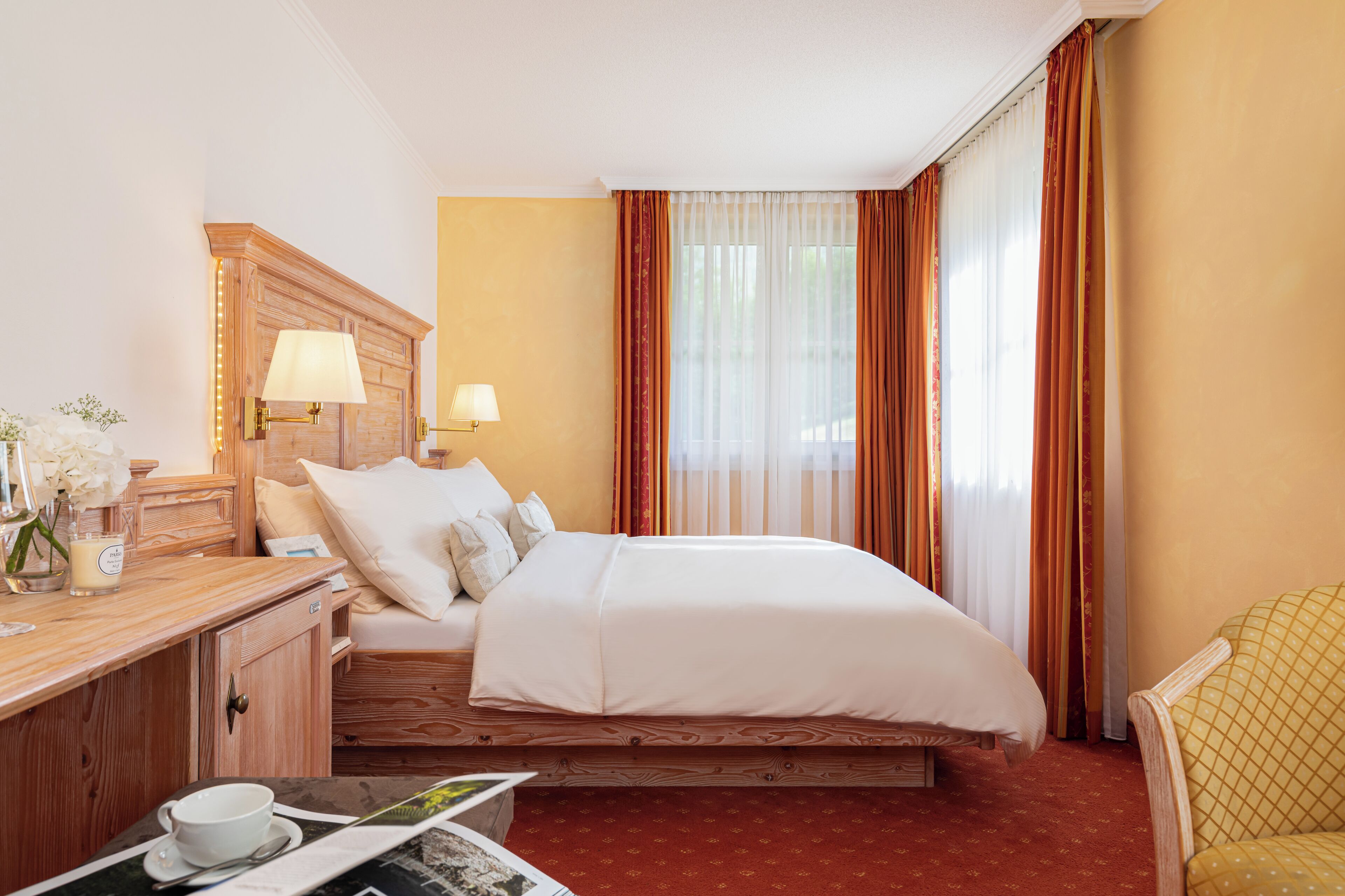 A hotel room with a bed, bedside lamps, a wooden headboard, a desk with a lamp, and a chair.