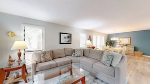 A living room with a sectional sofa, a glass coffee table, a side table with a lamp, and a dining area with a table and chairs.