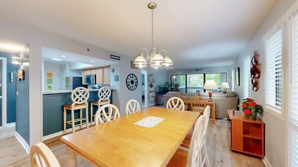 A modern kitchen with white cabinets, a dining area with a wooden table and chairs, and a living room with a sofa and a TV.