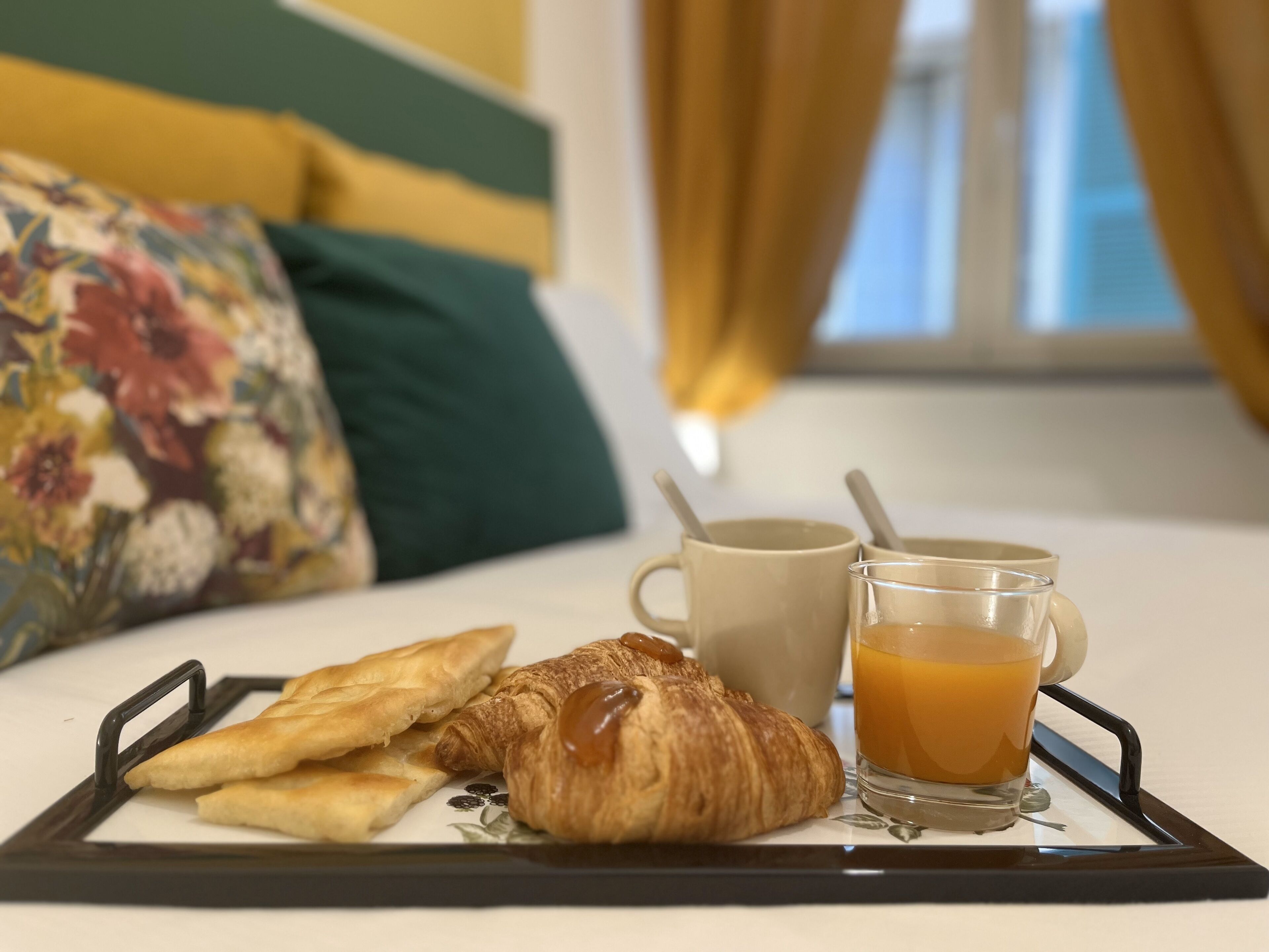 Bandeja de desayuno con pasteles y un vaso de zumo de naranja sobre una cama con almohadillas estampadas y vistas a una ventana con cortinas.