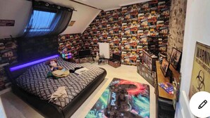 Ein Schlafzimmer mit schräg versiegelter Decke, einem Bett mit gemustertem Bettbezug, einer Spielkonsole auf dem Boden sowie einer Wand, die mit Büchern und Postern bedeckt ist.