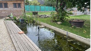Eine Holzterrasse neben einem Teich mit einem kleinen Wasserfall, umgeben von einer Rasenfläche und einer Holzhütte.