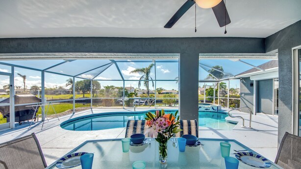 Une table de salle à manger au plateau en verre, agrémentée d’assiettes et de tasses bleues, placée devant une vue sur une piscine et un ciel dégagé.