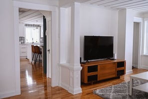 Uma sala de estar com uma TV sobre um suporte de madeira, uma área de jantar com cadeiras e uma cozinha ao fundo.