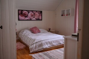 A bedroom with a bed, a pink pillow, a framed floral artwork, a small bedside table, and a pink curtain.