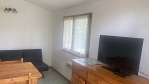 Et lille, godt oplyst rum med et træbord, to stole, en sort sofa, et fjernsyn på et træskab og et vindue med lette gardiner.