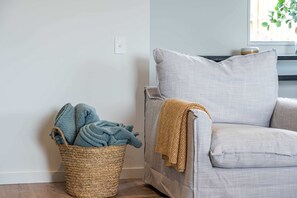 A corner of a room with a grey sofa, a woven basket with blue towels, and a window with a plant.