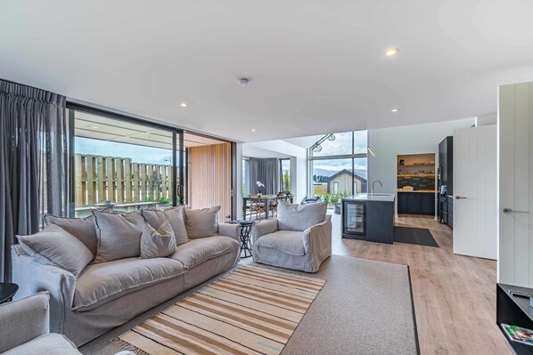 A modern living room with a sofa and armchairs, a kitchen area, and large windows offering a view of the outdoors.