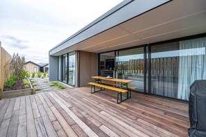 A modern outdoor patio with wooden decking, a wooden picnic table, and a glass-enclosed dining area.