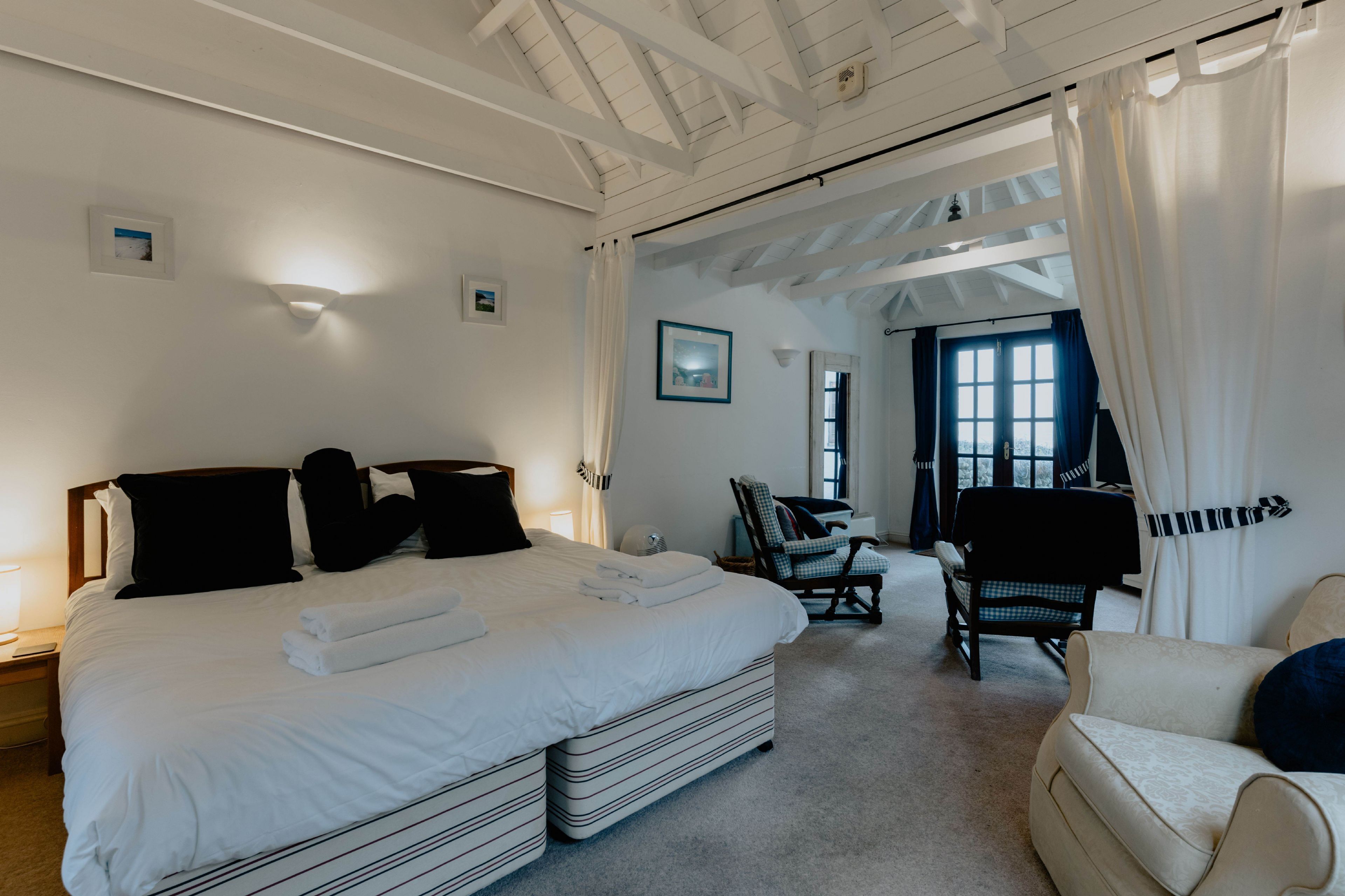 A hotel room with a large bed, a chair, and a sofa. There is a window with curtains, a picture on the wall, and a lamp on the nightstand.