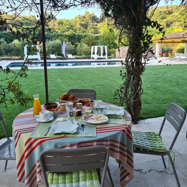 Un espace extérieur aménagé pour les repas, avec une table dressée pour deux personnes, une piscine et un kiosque en arrière-plan.