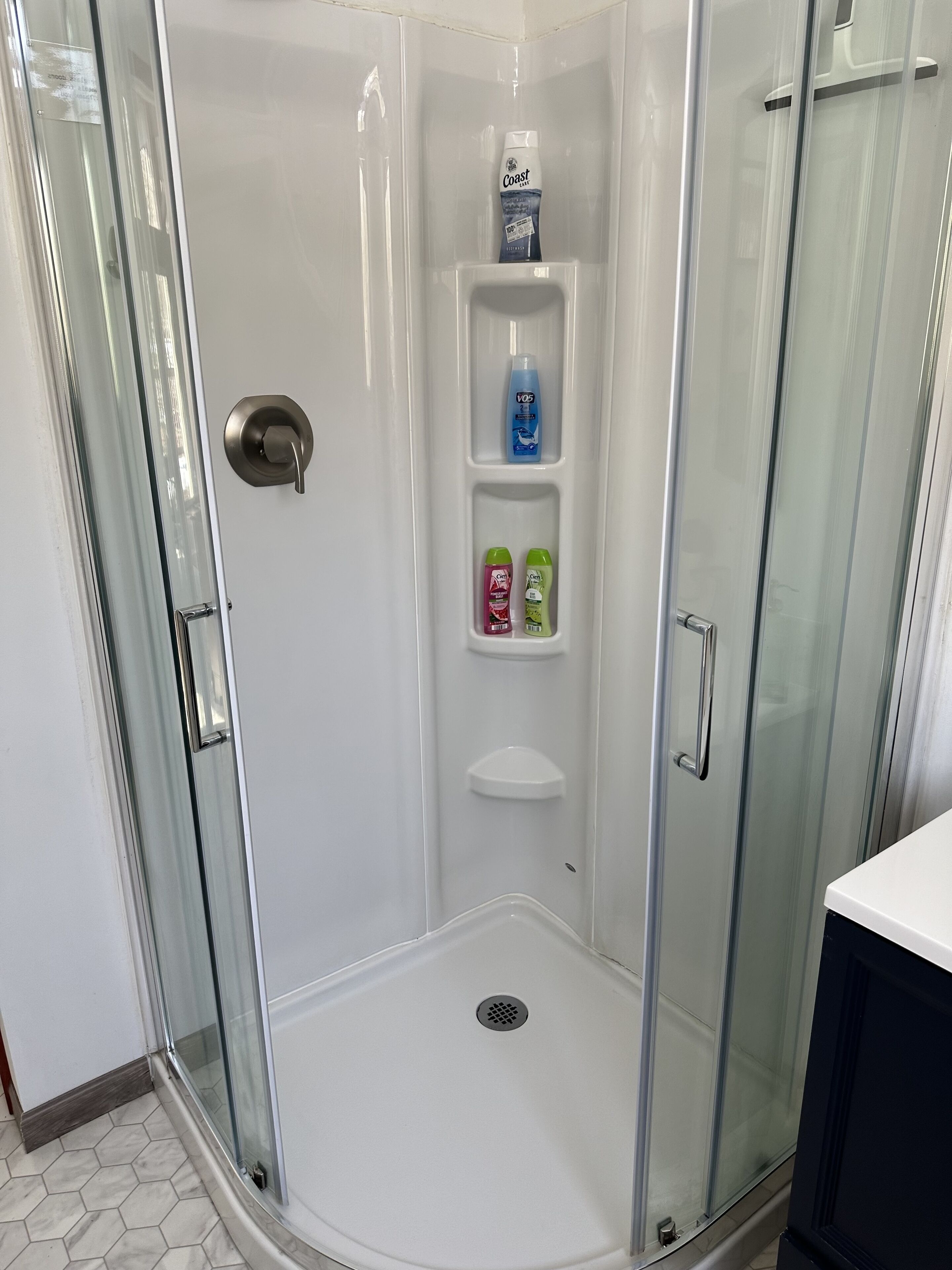 Eine Duschkabine mit Glastüren, ein weiß gefliester Boden, eine Handstange und ein Ablagefach mit Toilettenartikeln.