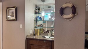 A bathroom with a wooden vanity, a glass shelf with decorative items, a framed picture, and a wall-mounted towel rack. - SUNNY SIDE UNIT 210  (South Padre Island)