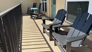 A covered outdoor area with two Adirondack chairs and a table, a door, and a window with blinds. - SUNNY SIDE UNIT 210  (South Padre Island)