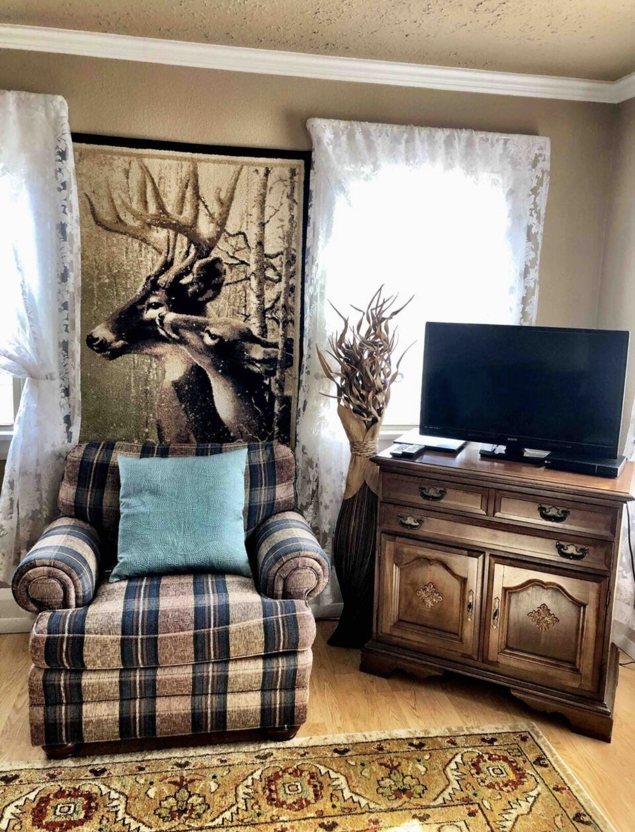 Een woonkamer met een geruite bank, een televisie op een houten kast en een groot hert schilderij aan de muur.